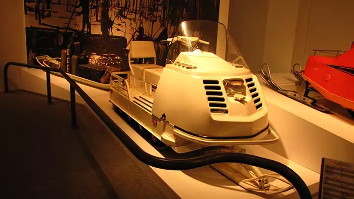Yellow snowmobile towing a sleight in a display case next to an orange snowmobile. 