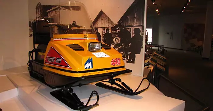 Yellow snowmobile with a single headlight on the hood. Photos on the wall behind. 