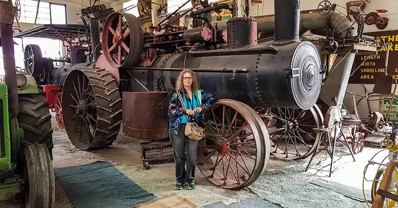 Linda Aksomitis standing beside a vintage steam engine tractor in a building housing agricultural equipment. 
